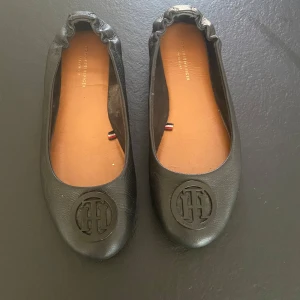 Svarta ballerinaskor från Tommy Hilfiger - Säljer ett par snygga svarta ballerinaskor från Tommy Hilfiger i nyskick. De är gjorda i skinn och har en rund tå med det klassiska TH-emblemet på framsidan. Perfekta för både vardag och fest!