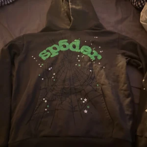 Svart hoodie från Sp5der - Säljer en svart hoodie från Sp5der med ett coolt spindelnätsmönster och gröna bokstäver. Den har en klassisk passform med långa ärmar och en bekväm huva. Perfekt för höst och vinter! Tyvärr inte äkta!