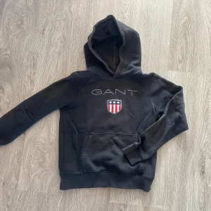 Marinblå hoodie från GANT - Säljer en snygg svart hoodie från GANT med deras logga och ett coolt emblem på framsidan. Använt skick därav priset. Den har en klassisk känguruficka och är perfekt för både vardag och chill. Passar för åldrarna 11-12 år.