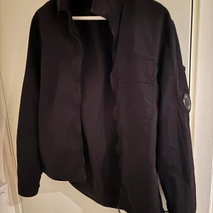 Svart jacka från C.P. Company - Säljer en stilren svart jacka från C.P. Company. Den har en cool detalj med en lins  på ärmen och är perfekt för höst och vår. långärmad och har en dragkedja framtill. Passar perfekt för en casual look! Skick 9,5/10 använd fåtal gånger 