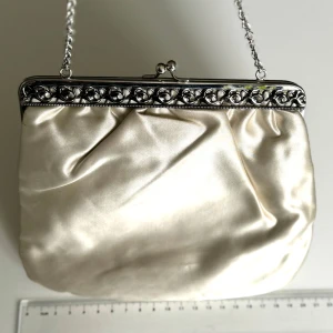Vintage vit kuvertväska med kedja - Vintage vit kuvert/handväska i satin med en elegant silverkedja. Väskan har en  metallram med dekorativa detaljer och praktisk knäppning upptill. Perfekt för festliga tillfällen som student, bröllop tex. Hämtas på Hägerstensåsen eller ök 🤍👜✨