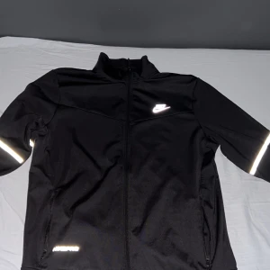 Svart träningsjacka från Nike - Säljer en svart träningsjacka från Nike med dragkedja och reflexdetaljer. Jackan har långärmade ärmar och en hög krage. Perfekt för löprundan eller gymmet.