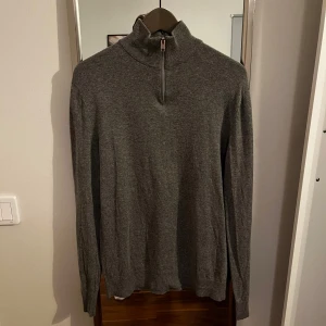 Grå tröja med dragkedja från Jack & Jones - Säljer en stilren grå tröja från Jack & Jones med dragkedja vid halsen. Tröjan har långa ärmar och är perfekt för kyligare dagar. Passar bra till både jeans och chinos. Hål vid nacken inget man tänker på med därav pris.