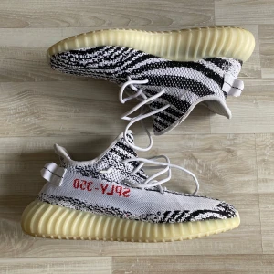 Adidas Yeezy Boost 350 V2 Zebra - Säljer ett par Adidas Yeezy Boost 350 V2 precis som originalet  i den ikoniska Zebra-färgen. Skorna har en svart och vit randig design med röd text på sidan. De har en bekväm och responsiv sula samt snörning för perfekt passform.