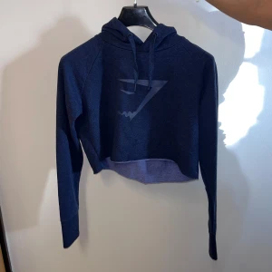 Mörkblå croppad hoodie från Gymshark - Säljer en snygg mörkblå croppad hoodie från Gymshark. Den har en stilren design med en diskret logga på framsidan och justerbar huva med dragsko. Perfekt för en avslappnad look.