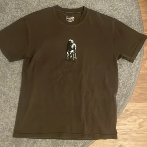 Brun t-shirt från Polar Skate Co. - Säljer en brun t-shirt från Polar Skate Co. med ett tryck av en person som sitter på en stol. Perfekt för en avslappnad stil. T-shirten har en normal passform och korta ärmar.