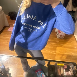 Blå sweatshirt med tryck - Säljer en blå sweatshirt med tryck 'Les Ateliers' i vitt på framsidan. Tröjan har långa ärmar och en avslappnad passform, perfekt för en casual look. Den är gjord i ett mjukt material som känns skönt mot huden.