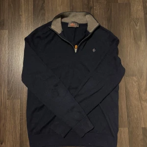 Mörkblå tröja med dragkedja från Morris - Säljer en stilren mörkblå tröja från Morris i 100% merinoull. Tröjan har en dragkedja framtill med en snygg läderdetalj och en kontrasterande krage i grått. Perfekt för kyligare dagar. Storlek L men passar som M.