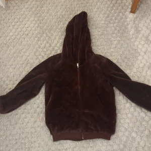 Vinröd teddyjacka från Rainbow - Mysig vinröd teddyjacka med huva från Rainbow. Jackan har dragkedja framtill och långa ärmar. Perfekt för kyliga dagar när du vill hålla dig varm och bekväm.