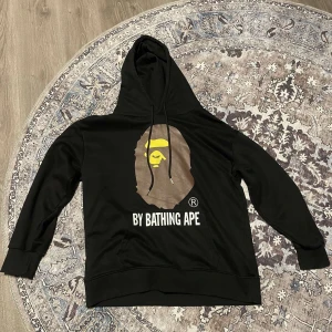 Svart hoodie från  Bathing Ape - Säljer en svart hoodie från A Bathing Ape med det ikoniska aptrycket på framsidan. Hoodien har en justerbar huva med svarta snören och ribbade muddar vid ärmslut och nederkant. Perfekt för en stilren och avslappnad look.