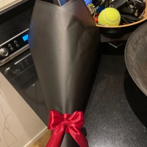 Bukett med röda rosor i svart omslag - En elegant bukett med konstgjorda röda rosor inslagna i ett stilrent svart omslag. Perfekt som en romantisk gest eller dekoration. Rosorna är gjorda av tyg och har en vacker glans som ger ett realistiskt utseende. Omslaget är prytt med ett rött band för en extra touch av elegans.