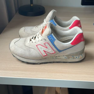 Beige sneakers från New Balance - Snygga beige sneakers från New Balance med röda och blå detaljer. Skorna har snörning och en bekväm sula med färgglada prickar. Perfekta för en avslappnad stil.