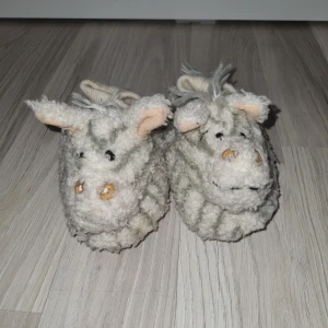 Gråa tofflor med djurmotiv - Mysiga gråa tofflor med ett sött djurmotiv. De har ett randigt mönster och är tillverkade i ett mjukt material. Perfekta för att hålla fötterna varma inomhus.