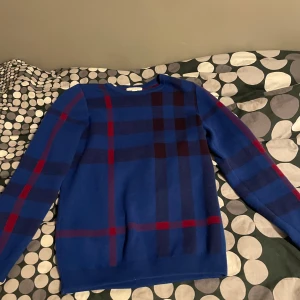 Snygg Burberry tröja i kashmir  - Snygg blå burberry tröja i kashmir med ett klassiskt rutmönster i rött och mörkblått. Tröjan har långa ärmar och en bekväm passform. Perfekt för en stilren look.
