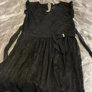 Svart spetsklänning med rosett - Elegant svart spetsklänning med en stor rosett dekorerad med pärlor i midjan. Klänningen har korta ärmar och en rund halsringning med pärldetaljer. Perfekt för en stilfull look.