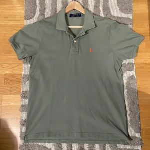 Ralph Lauren Piké  - Säljer en stilren grön pikétröja från Ralph Lauren med klassisk krage och knappar. Den har en broderad logotyp i orange på bröstet. Perfekt för en avslappnad men snygg look. Använt den ett tag och känner att det är dags och gå vidare! Vid frågor så ärt det bara och ställa!