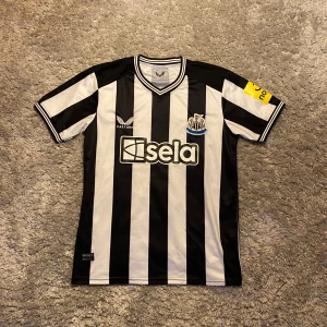 New castle fotbollströja - Snygg svartvit randig fotbollströja från Castore med klubbmärke och sponsorlogga på framsidan. Tröjan har korta ärmar och en klassisk V-ringning. Perfekt för fotbollsfans som vill visa sitt stöd.