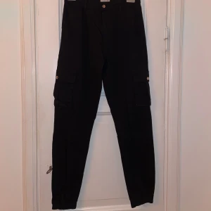 Svarta cargopants med fickor - Snygga svarta cargopants med flera praktiska fickor och knappdetaljer. Perfekta för en avslappnad stil med en touch av funktionalitet. De har en bekväm passform och elastiska muddar vid bensluten.