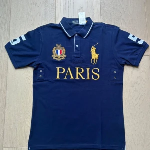 Mörkblå pikétröja med Paris-broderi - Är köpt begagnat och har använd i två månader efter det ungefär. den var nyskick när jag fick den och inga skador har tillkommit. Det står storlek M men passar nog bättre som storlek S. Bara att skicka om det är några frågor. 