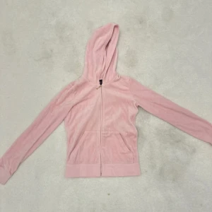 Rosa hoodie från Juicy Couture - Säljer en mjuk och gosig rosa hoodie från Juicy Couture. Den har en dragkedja framtill och en stor logga med krona på ryggen i leopardmönster. Perfekt för en avslappnad stil med en touch av glamour. Alla diamanter sitter kvar och av god kvalitet. Storleken är 14 år men kan även passa mindre tack vare muffarna