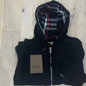 Svart huvtröja med dragkedja från Burberry - Snygg svart huvtröja från Burberry med dragkedja och klassiskt rutigt mönster i huvan. Perfekt för en stilren look. Tröjan har långa ärmar och ribbade muddar vid ärmslut och nederkant. Ett måste för garderoben!