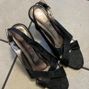 Svarta klackskor med remmar - Eleganta svarta klackskor från Neno med remmar och öppen tå. Skorna har en stilren design med ett diskret mönster och justerbart spänne vid ankeln. Perfekta för en kväll ute eller en speciell tillställning.