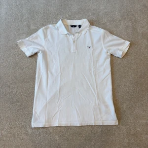 Vit pikétröja från gant  - Säljer en klassisk vit pikétröja från gant med krage och korta ärmar. Perfekt för en stilren och avslappnad look. Tröjan har en liten logga på bröstet och är tillverkad i ett bekvämt material.