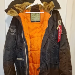 Svart jacka från Alpha Industries - Svart jacka från Alpha Industries med ett coolt orange foder. Jackan har en pälsfodrad huva och flera fickor med dragkedjor. Perfekt för kalla dagar! Garderobsrensning så kika gärna på övriga auktioner. 