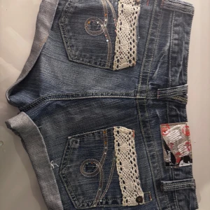 Jeansshorts med spetsdetaljer - Snygga jeansshorts med unika spetsdetaljer på fickorna. De har en cool design med dekorativa sömmar och knappar. Perfekta för en avslappnad stil med en touch av elegans.