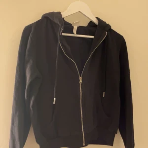 Svart hoodie från H&M - Säljer en svart hoodie från H&M med dragkedja och huva. Den har långa ärmar och snörning vid huvan. Perfekt för en avslappnad stil.