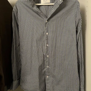 Rutig skjorta från The Shirt Factory - Snygg rutig skjorta från The Shirt Factory i blått och vitt. Skjortan har en slim fit passform och är strykfri, perfekt för en stilren look. Den har långa ärmar och knappar framtill. Den är nästan i nyskick och riktigt stekig