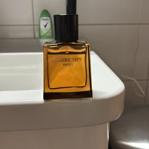 Burberry Hero Eau de Parfum - Säljer en elegant Burberry Hero Eau de Parfum (med box) i en stilren, fyrkantig flaska med en djup bärnstensfärg. Flaskan har en svart kork och ger en känsla av lyx och modernitet. Perfekt för den som vill ha en sofistikerad doftupplevelse. 