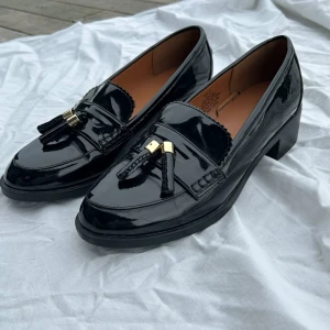 Svarta loafers från H&M - Snygga svarta loafers i lack från H&M med dekorativa tofsar och gulddetaljer. Perfekta för en stilren look.