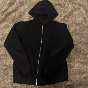 Svart ribbad hoodie med dragkedja - Säljer en stilren svart hoodie med dragkedja och huva. Den har en ribbad textur som ger en cool look. Perfekt för en avslappnad stil. Den är stickad och man sticker inte ut med den den har inga stora skador och perfekt för hösten som kommer runt hörnet 