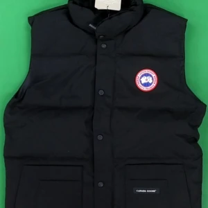 Svart dunväst från Canada Goose - Säljer en svart dunväst från Canada Goose med ett broderat märke på bröstet. Västen har knappar framtill och två praktiska fickor. Perfekt för kyligare dagar.