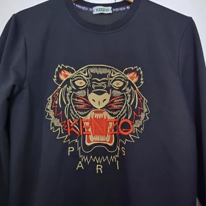 Svart tröja från Kenzo - Snygg svart tröja från Kenzo med ett iögonfallande tigerbroderi i orange och beige på framsidan. Tröjan har långa ärmar och rund halsringning. Perfekt för att ge din outfit en cool touch!