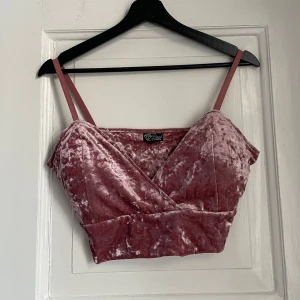 Rosa sammetstopp med smala axelband - Säljer en snygg rosa sammetstopp med smala axelband och omlott-design. Perfekt för en trendig look. Toppen är croppad och har en mjuk, glansig yta som ger en lyxig känsla.