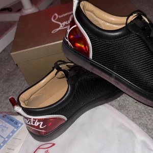 Christian Louboutin Skor - Snygga svarta sneakers från Christian Louboutin med nitar på tån och en glansig röd häl med vit text. Skorna är använda en gång och säljer därför att dom inte passade mig. Extra spikar, box, dustbag medföljer. Bara att höra av er om ni har frågor eller funderingar :)