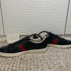 Svarta   Gucci ace  - Snygga svarta sneakers från Gucci med klassiska gröna och röda ränder på sidan. Skorna har snörning och en präglad logotyp på ytan. Perfekta för en stilren look. Fint skick köp elle byt mot nå kul 