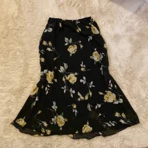 Svart blommig kjol från S. Maria - Snygg svart kjol med vackra gula blommor från S. Maria. Kjolen har en elastisk midja för bekväm passform och är perfekt för en stilfull look. Den är i en längre modell och har ett flytande fall.