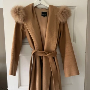 Beige kappa med pälskrage från Hollies - Elegant beige kappa från Hollies med lyxig pälskrage och knytbälte i midjan. Perfekt för kyliga dagar med sin stilrena design och långa ärmar. Inköpt för 6500kr.