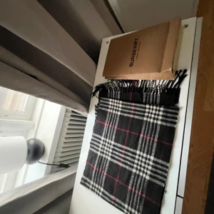 Rutig halsduk från Burberry - Säljer en klassisk rutig halsduk från Burberry i 100% lammull. Den har en stilren design med svarta, vita och röda rutor. Perfekt för att hålla dig varm och snygg under kyliga dagar. Kommer med originalförpackning.
