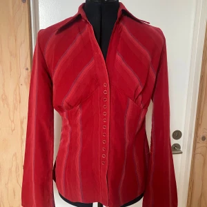 Röd randig skjorta - Snygg röd skjorta med vertikala ränder och knappar framtill. Skjortan har långa ärmar och en klassisk krage. Perfekt för en stilren look. Köptes secondhand i Köpenhamn för något år sen. 