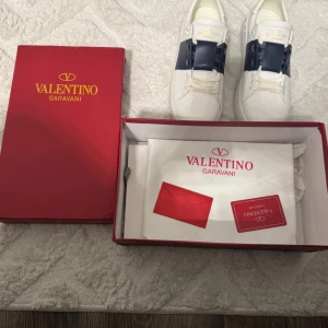 Valentino Garavani sneakers i vitt och blått - Snygga Valentino Garavani sneakers i vitt med en bred blå rem över snörningen. Skorna har en stilren design med en vit sula och subtila detaljer. Perfekta för en trendig look.