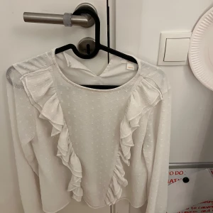 Vit blus med volangdetaljer - Säljer en söt vit blus med volangdetaljer framtill. Blusen har ett diskret prickigt mönster och långa ärmar. Perfekt för en stilren look. Den är gråt gina strl 158-164 men skulle säga xs. Skriv om frågor osv💗 priset går att diskutera