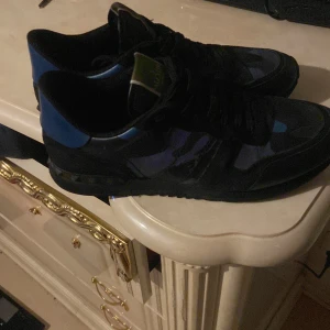 Valentino rockrunners  - Snygga svarta sneakers med blå detaljer från Valentino. Skorna har ett coolt camouflage-mönster och klassisk snörning. Perfekta för en stilren look.