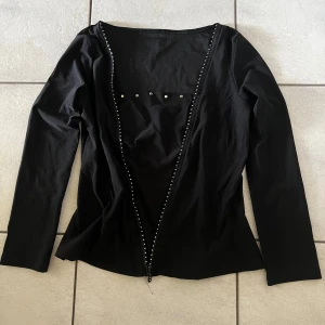 Svart långärmad topp med dragkedja - Snygg svart långärmad topp med dragkedja framtill och dekorativa pärlor längs kanten. Perfekt för en stilren look.