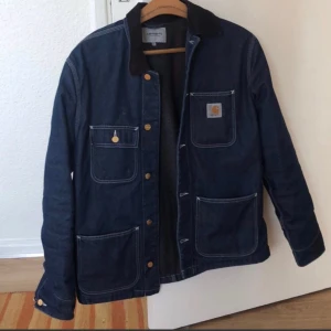 Blå jeansjacka från Carhartt - Säljer en snygg blå jeansjacka från Carhartt med svarta detaljer. Jackan har flera praktiska fickor och knappar framtill. Perfekt för en avslappnad stil. Som ny, storlek L