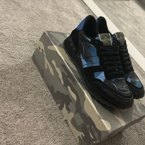 Valentino Rockrunner  - Snygga Valentino Rockrunner sneakers i svart och blått med kamouflagemönster. Skorna har en klassisk snörning och är tillverkade i en kombination av skinn och syntetmaterial. Perfekta för en trendig look.