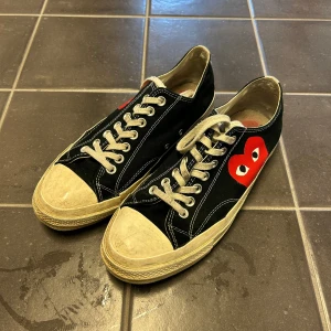 Svarta converce CDG - Säljer ett par svarta Converse sneakers med en röd hjärtdetalj på sidan. Skorna har vita snören och en klassisk gummisula. Perfekta för en avslappnad stil.